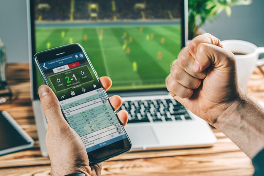 A hand holding a mobile showing footbal odds while a football match game is being streamed via laptop.