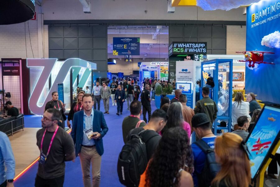 Los asistentes caminan por un pasillo alfombrado de azul en la convención SiGMA South America 2026. Se pueden ver los modernos stands de BGAMING y Xtremepush, y en el techo de una gran sala cuelgan carteles indicativos que señalan el Pabellón E.