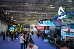 Una vista panorámica del bullicioso pabellón de exposiciones de SiGMA South America 2026. La escena muestra los amplios y modernos stands de Altenar y Spribe, con luminosas pantallas digitales. Los asistentes deambulan por los pasillos, cubiertos de moqueta azul, bajo un alto techo industrial.