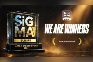 A celebratory award graphic set against a textured, dark gold and black background with a dramatic spotlight effect from the top right. On the left side, a large 3D glass and metallic trophy is prominently featured; it showcases the chrome ‘SiGMA’ logo over a gold ‘EUROPE’ plate, resting on a black tier inscribed with ‘BEST E-SPORT SUPPLIER 2026’ in gold lettering. On the right, the main headline reads ‘WE ARE WINNERS’ in bold white capital letters with a slight green glow. Above the headline sits the square white ‘SiGMA AWARDS EUROPE’ logo, while below it, the category text ‘BEST E-SPORTS SUPPLIER’ is displayed between two small golden laurel wreath icons.