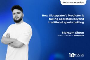 A promotional graphic for an interview from Focus Gaming News set against a textured blue gradient background. On the left, a black-and-white portrait shows Maksym Shtun standing with his arms crossed, wearing a white t-shirt. On the right, the main headline reads: ‘How Slotegrator's Predictor is taking operators beyond traditional sports betting’ in bold white lettering. Below the headline, Shtun’s name and job title, ‘Product Owner at Slotegrator’, are displayed. A white pill-shaped button containing the text ‘Exclusive Interview’ is located in the top-right corner, and the Focus Gaming News logo is positioned in the bottom-right corner.