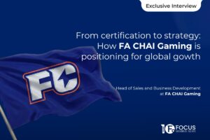 A promotional graphic for an exclusive interview titled ‘From Certification to Strategy: How FA CHAI Gaming Is Positioning for Global Growth'. The design features a 3D-styled hand holding a flagpole with a waving navy blue flag that bears the ‘FC’ logo. The background is a blue gradient with a subtle silhouette of a map of South America. The Focus Gaming News logo is positioned in the bottom-right corner.