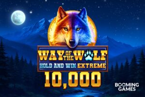 A promotional title graphic for the online slot game ‘WAY OF THE WOLF HOLD AND WIN EXTREME 10,000’ by Booming Games. The background depicts a nocturnal wilderness scene with a pine forest, mountains under a starry sky, and a bright full moon on the left. In the upper centre, a large portrait of a wolf's head is framed by a golden coin; the wolf is dramatically lit, with the left side of its face glowing in warm orange and the right side in cool blue. Below the wolf, the game’s logo is displayed on a wooden-textured banner with gold and blue lettering, featuring a glowing blue paw print inside the word ‘WOLF’. The number ‘10,000’ is written underneath in large, bold gold 3D numbers, and the white Booming Games logo is positioned in the bottom-right corner.