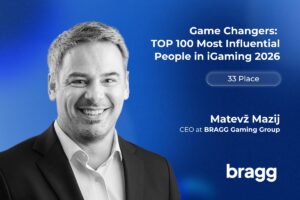 A promotional feature graphic set against a textured, deep blue background with a subtle light glow. On the left side, a black-and-white portrait features a smiling Matevž Mazij wearing a dark suit jacket over a light-coloured collared shirt. In the upper right, bold white text reads: ‘Game Changers: TOP 100 Most Influential People in iGaming 2026’. Below this headline, a translucent white pill-shaped outline contains the text ‘33 Place’. Mazij’s name and job title, ‘CEO at BRAGG Gaming Group’, are printed in white towards the bottom right, positioned just above the white BRAGG company logo in the bottom-right corner.