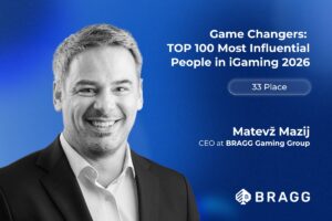 A promotional feature graphic set against a textured, deep blue background with a subtle light glow. On the left side, a black-and-white portrait features a smiling Matevž Mazij wearing a dark suit jacket over a light-coloured collared shirt. In the upper right, bold white text reads: ‘Game Changers: TOP 100 Most Influential People in iGaming 2026’. Below this headline, a translucent white pill-shaped outline contains the text ‘33 Place’. Mazij’s name and job title, ‘CEO at BRAGG Gaming Group’, are printed in white towards the bottom right, positioned just above the white BRAGG company logo in the bottom-right corner.