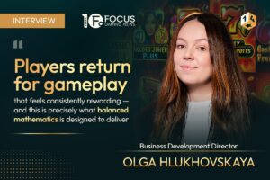 A promotional interview graphic set. On the right, a colour portrait shows Olga Hlukhovskaya with long auburn hair smiling slightly, positioned below a 1x2 gaming emblem in the top-right corner. In the top-left corner, a gold rectangular badge reads ‘INTERVIEW’, next to the Focus Gaming News logo. On the left, a large quote in gold and white text reads: “Players return for gameplay that feels consistently rewarding — and this is precisely what balanced mathematics is designed to deliver”. In the bottom-right corner, her title ‘Business Development Director’ appears above her name, ‘OLGA HLUKHOVSKAYA’, written in bold gold capital letters.