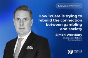 An interview graphic set against a blue background. On the left, a black and white portrait of Simon Westbury, Chairperson of the 1xCare Advisory Board. To the right, text reads: 'Exclusive Interview', followed by 'How 1xCare is trying to rebuild the connection between gambling and society', and 'Simon Westbury, Chairperson 1xCare Advisory Board'. The 'Focus Gaming News' logo is in the bottom right corner.