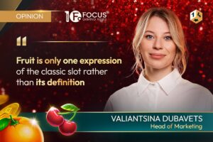A promotional opinion feature graphic set. On the right, a colour portrait shows Valiantsina Dubavets, positioned below a '1x2' diamond-shaped gaming emblem in the top-right corner. In the top-left corner, a gold rectangular badge displays ‘OPINION’, positioned next to the Focus Gaming News logo. On the left side, a quote in gold and white text reads: “Fruit is only one expression of the classic slot rather than its definition”. In the bottom-right corner, her name ‘VALIANTSINA DUBAVETS’ is printed in bold white capital letters over a teal banner line, with her title ‘Head of Marketing’ written in gold underneath.